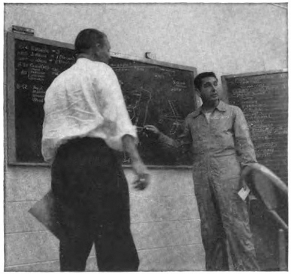 Two men stading at a blackboard, one with his back to the camera