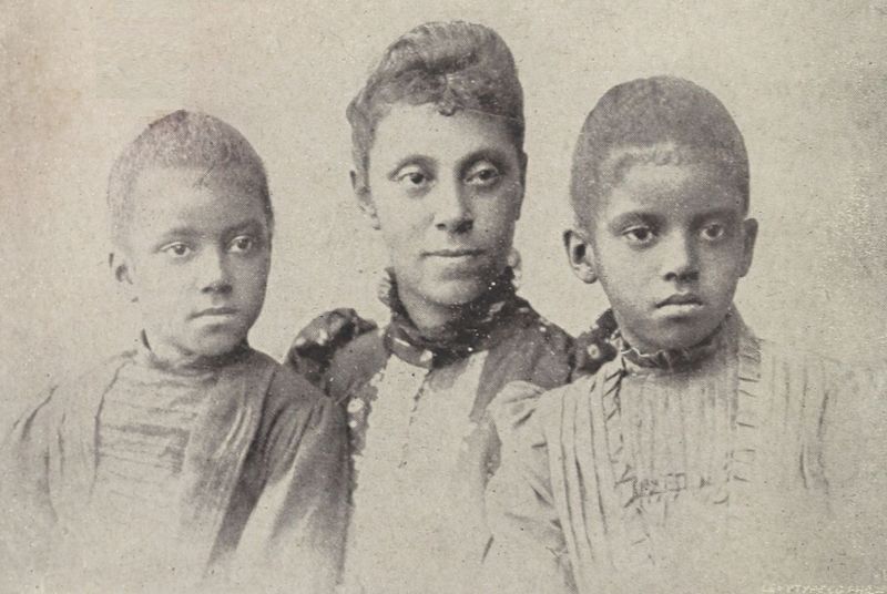 The illustration shows Mrs. N. F. Mossell (Gertrude Bustill Mossell) with her daughters, Florence and Mary, standing on either side of her. Mossell’s hair is styled in an updo, while her daughters have short hair. All three wear Victorian dresses decorated with pleats, ruffles, and buttons.