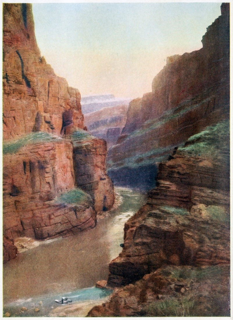 Color photograph with canyon walls extending up the sides; the left side is sunlit, the right in shadows; a brown river curls through the bottom of the canyon; a light blue sky forms a V at the top between the two canyon walls.