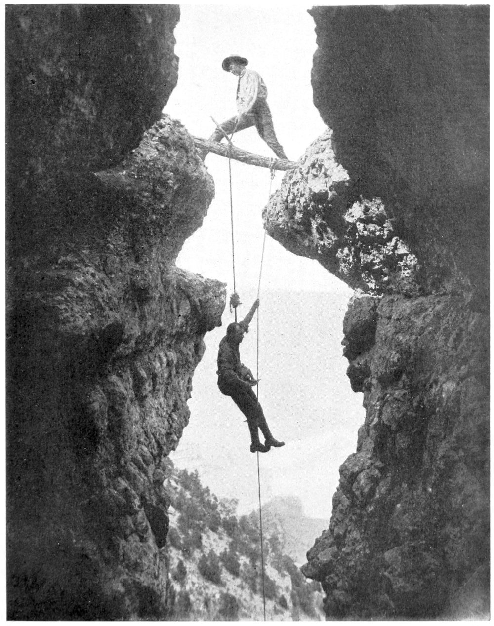 Emery is suspended in a narrow canyon by a rope, holding onto another rope, the bottom of which is not visible. Both ropes are affixed to a log that spans the gap between the walls of the canyon. Ellsworth straddles the gap with one foot at each end of the log.