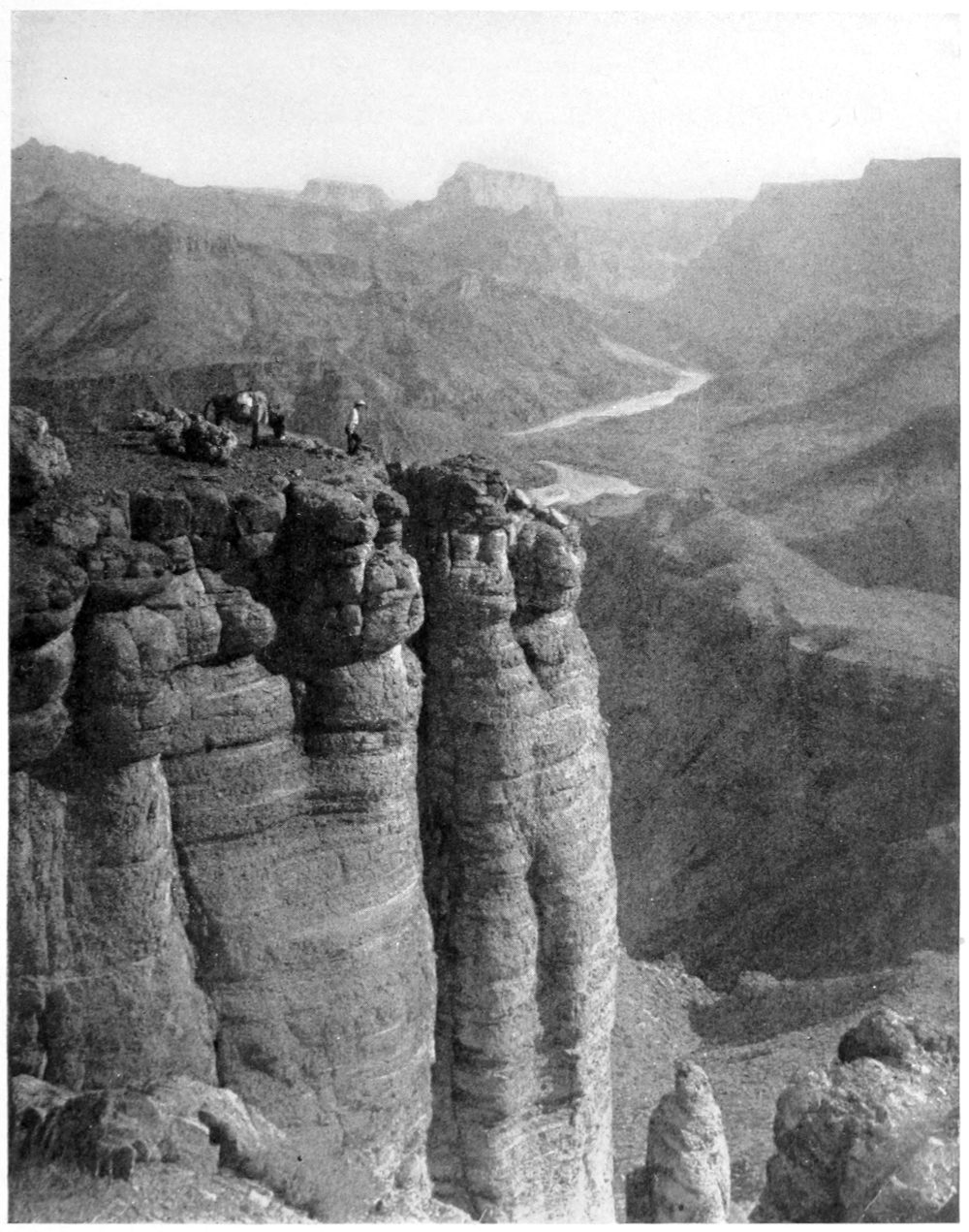 View of the canyon from atop a canyon wall. A man and a burro are on top of the near canyon ridge. The river snakes through an open section of the canyon in the distance.
