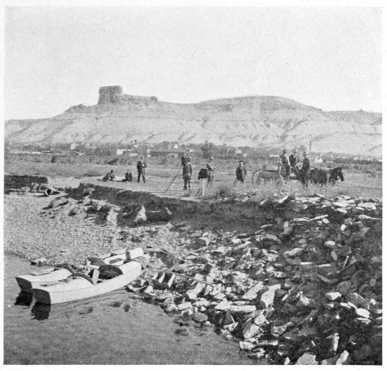 Two rowboats and gear are beached on a rocky river shore. A little above on a flat section, two mean are seated, four are standing; two cameras are on tripods. Two men are riding in a horse-drawn cart, and another man watches on horseback. A village is in the background, behind which is a high, tree-less mesa.
