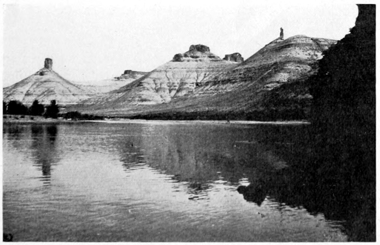 Calm waters are in the foreground. On the opposite shore are conical, eroded hills capped by uneroded blocks. The erosion has made stripes visible of various geologic strata.