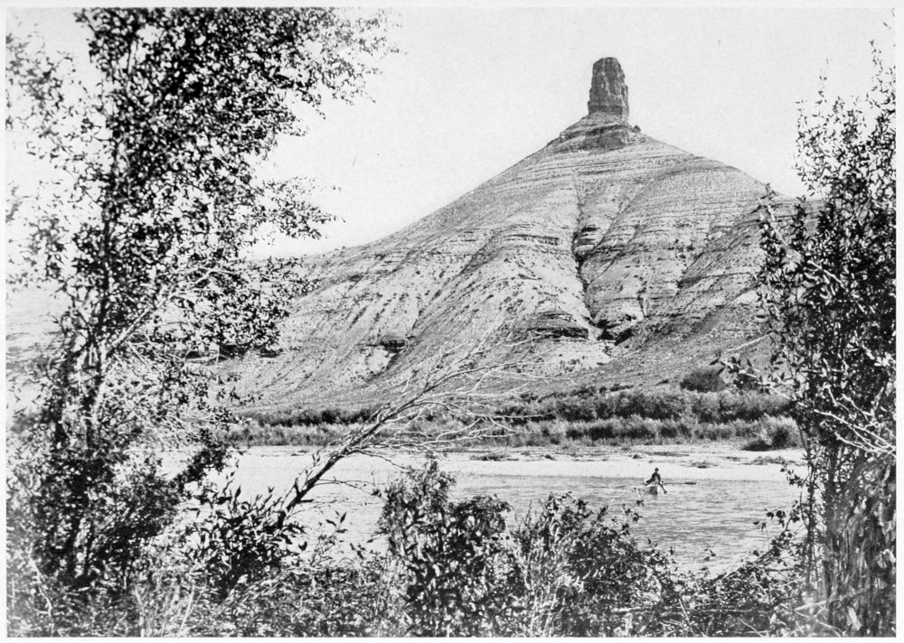 View of a high hill of stratified rock, topped with a stone pointing upwards like a thumb. The river is in front of the rock, with shrubs and low vegetation in the foreground and along the sides of the photo.
