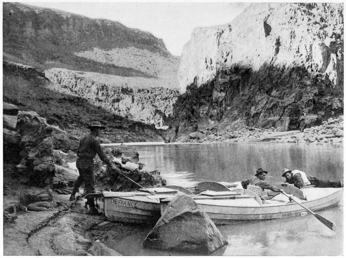 The author stands on the shore at left holding the 'Defiance' by a line at the stern. Emery is seated in the hatch and Luzon lies on the prow. The river is calm and reflects low canyon walls in the distance.