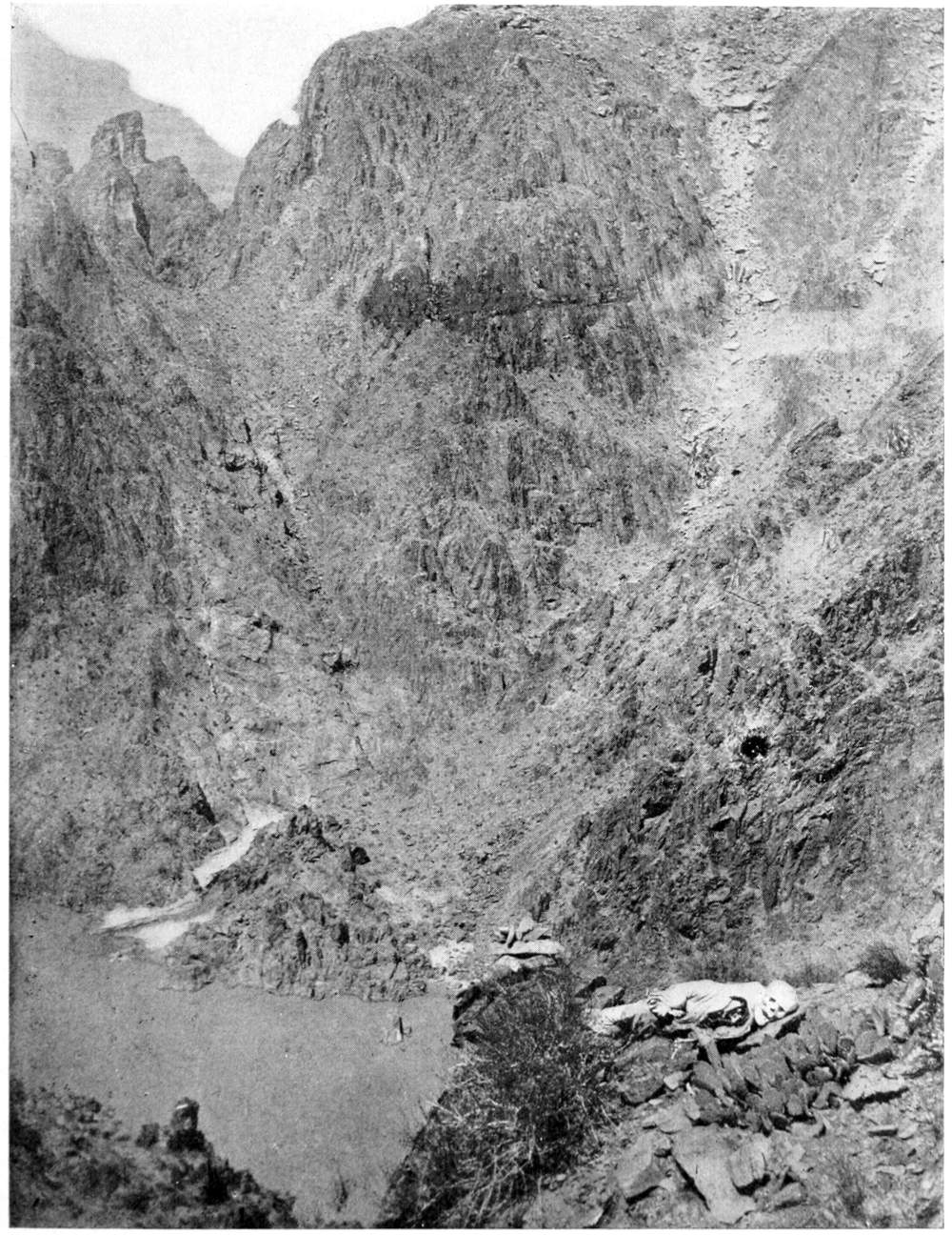 Steep, rugged canyon walls dominate the photo. In the lower right is a human skeleton lying on a ledge among cacti and rocks.