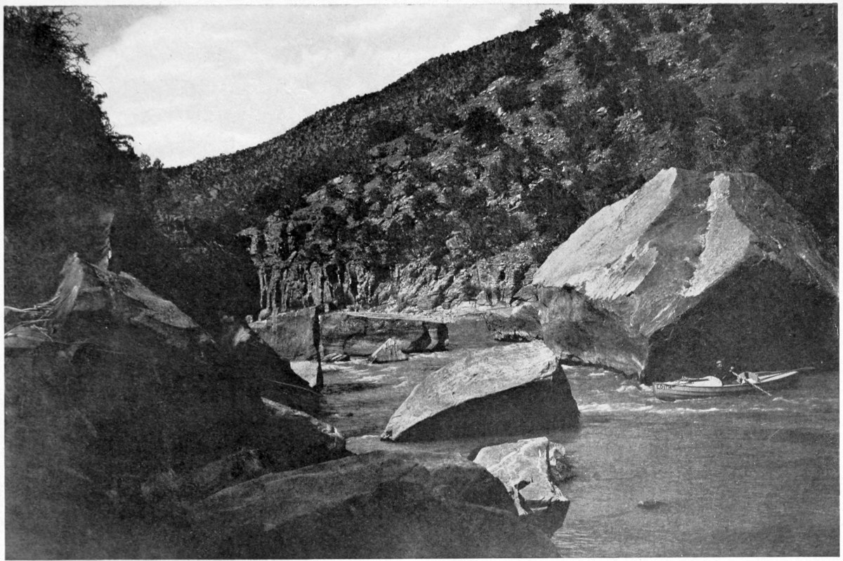 The 'Edith' is in the river in front of a boulder, about 10 times the height of the rowboat. Other large boulders are scattered through the river. The hills on the far side of the river are covered with trees.