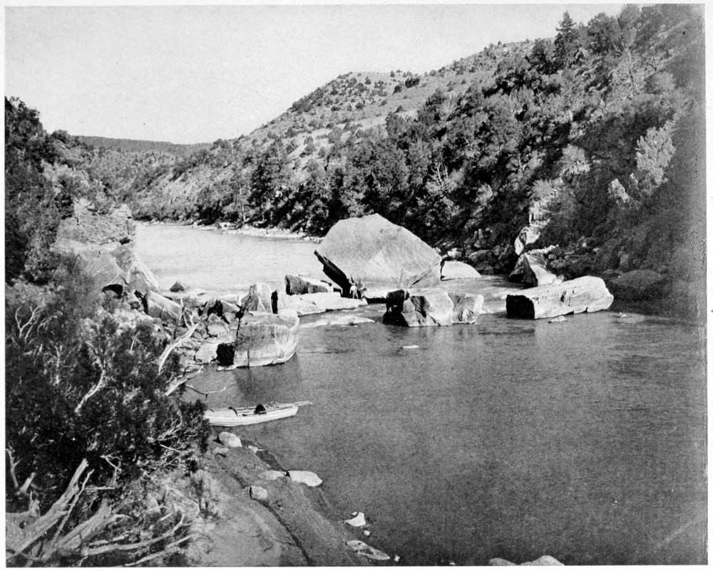 In the middle of the wide river are huge boulders across its entire width. One of the rowboats is grounded at the shoreline before the boulders. Two men are standing on a flat rock in the center of the river. Gentle hills on both sides of the river are covered with trees and shrubs.