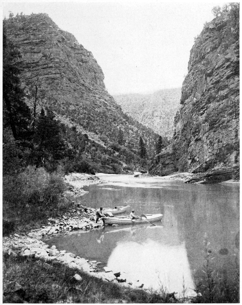 Both rowboats are beached on the left shore. One man holds onto a boat with another man in the cockpit. The water is calm and relfects the low canyon walls beyond the boats.