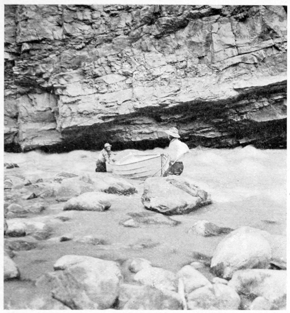 Two men, up to their knees in the river, portage a boat through a rocky section; the river roils next to the canyon wall.