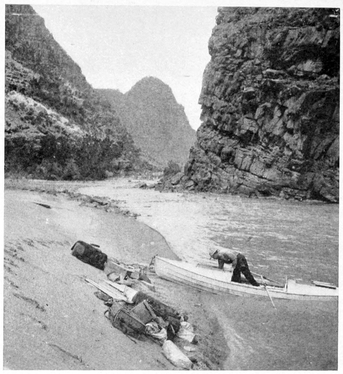 A boat is beached on a sandy shoreline. A man in the cockpit reaches into the interior of the boat. Piles of gear are on the shore.