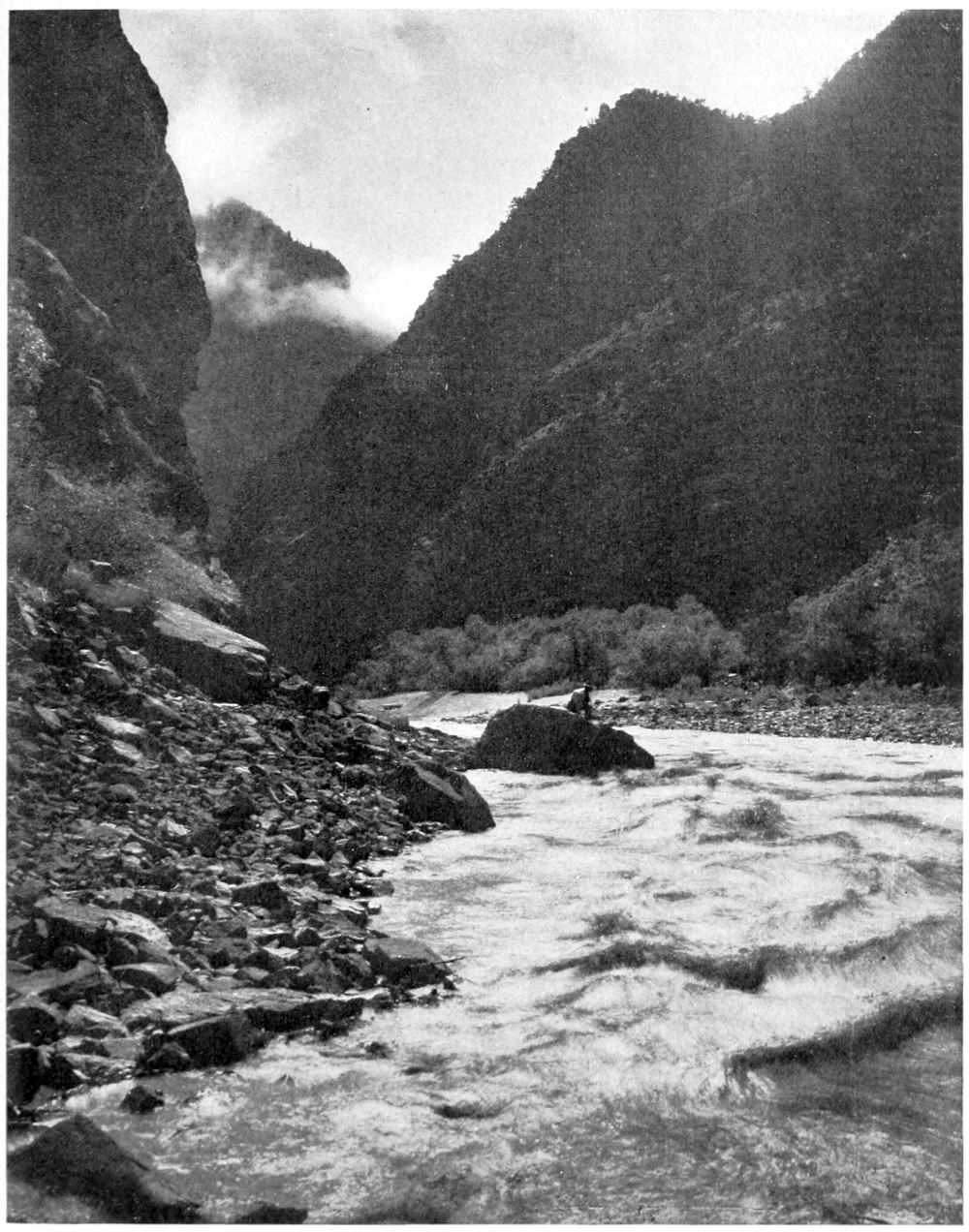 A man sits on a large rock that extends from the shore into the churning river water. The upper canyon walls are obscured by clouds.