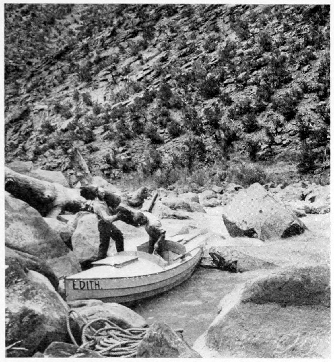 The 'Edith' is beached among large rocks on the shore. One man passes two bags of gear from the hatch to another man standing among the rocks.