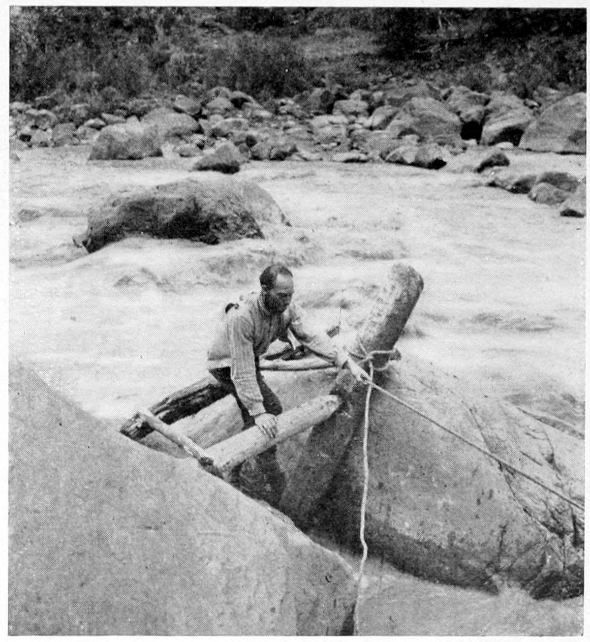 A man stands between two large boulders in the middle of raging water. He holds a rope with his left hand that is tied to an upright log; his right hand rests on a smaller horizontal log wedged between a boulder and the upright log.