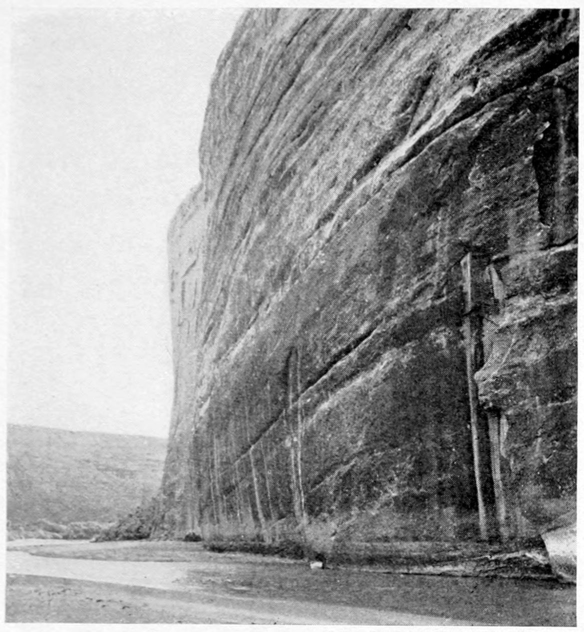 A calm river curves around a massive, vertical, nearly smooth canyon wall on the right. Low hills are in the distance on the left.