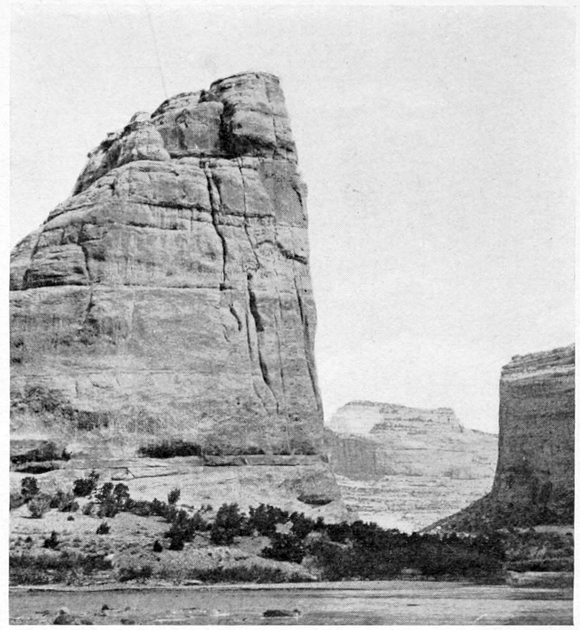 A smooth river flows across the bottom of the photo. A flat-topped mesa is on the right. In the center, a canyon wall rises several hundred feet, sloping downward towards the left side of the photo.