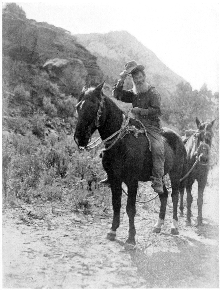 A man on horseback raises his hat to the photographer. A burro is tied by a leash behind his horse. Hills rise up behind him to the horizon.