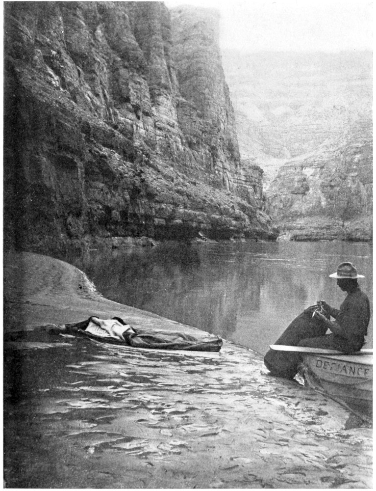 At the side of the river, a man sits on an oar at the prow of the 'Defiance' with a duffle bag. On the shore next to the boat is bedding yet to be rolled and stowed. Beyond, canyon walls rise beyond the top of the photo.