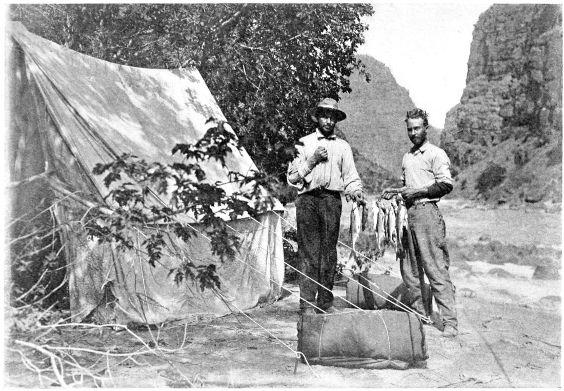 The Kolb brothers stand ashore a few feet apart, holding a line of fish between them. A tent is on the left, baggage is on the ground around the men, and the river flows behind them.
