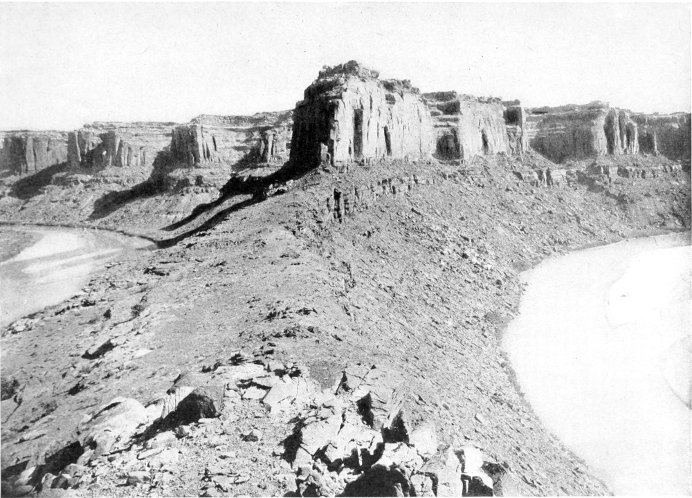 The river curves into the photo on both the right and left sides. The photo was taken on the spit of land between the two curves. Beyond the curves rise three rock formations that cast similar shadows on the land below them.