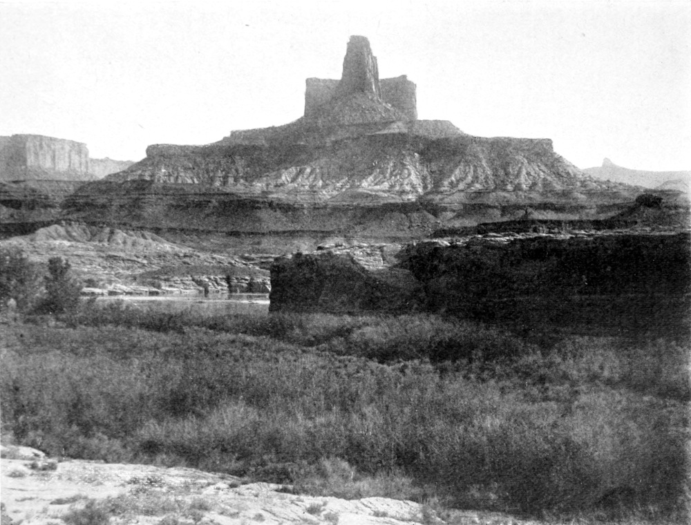 A large mesa is beyond one shore of the river. Atop the mesa in the center is a rock formation that extends towards the sky. It is in front of a shorter rectangular formation giving the appearance of the top half of a cross.