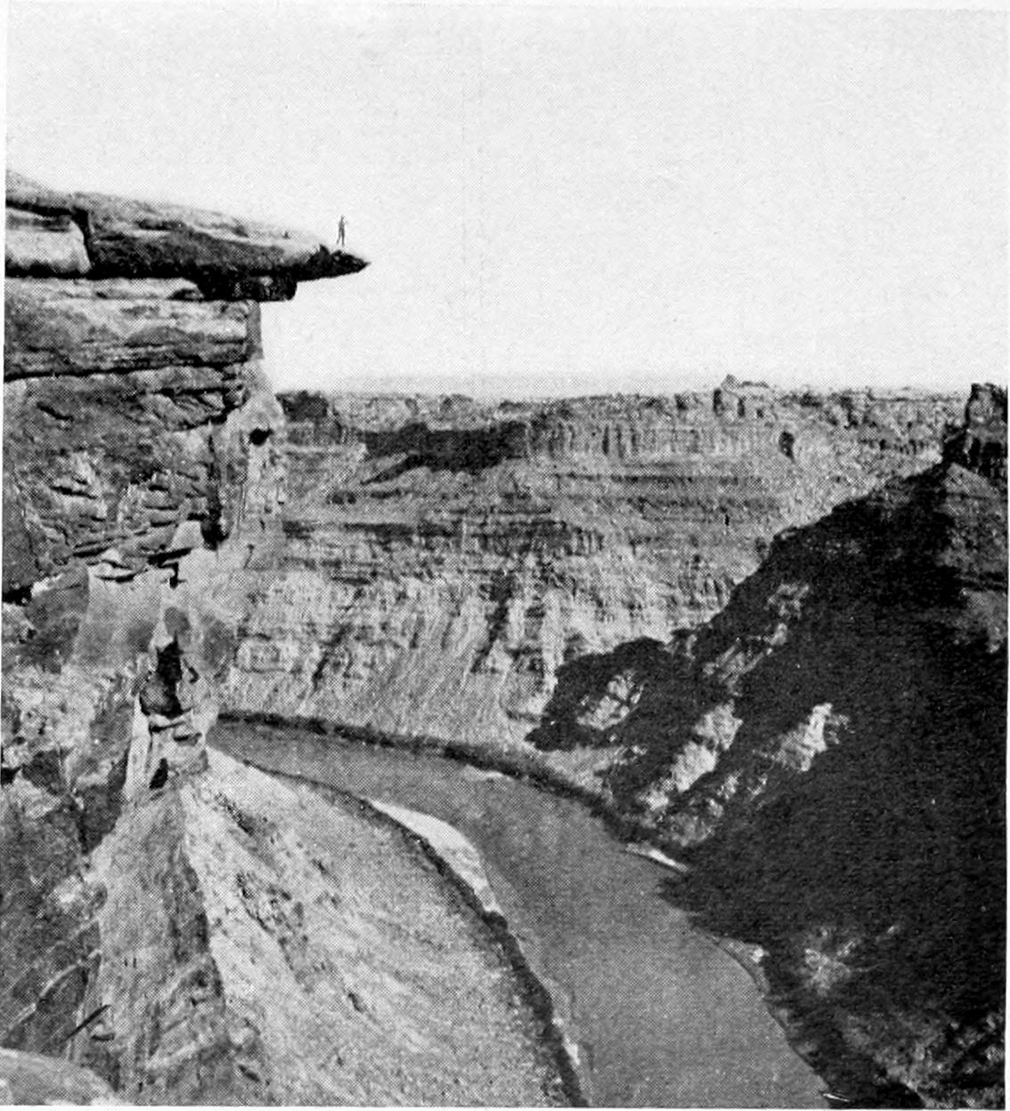 The river curves from the lower right to the left of the photo. Canyon walls extend upwards on both sides of the river. On the left, an outcrop of rock extends over the canyon wall. A tiny figure of a man is atop the outcrop.