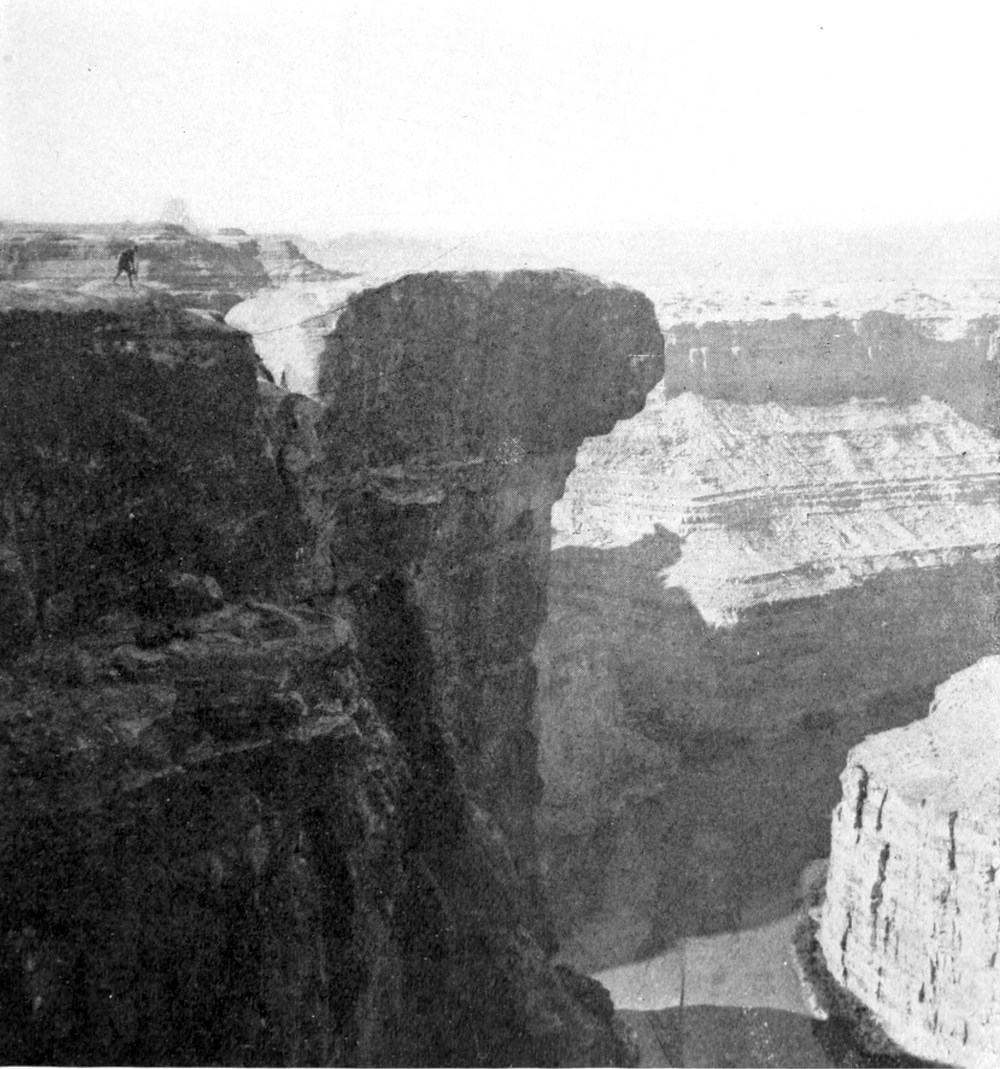 A tall canyon wall in the shadows dominates the left half of the photo. A man stands on top of the wall. On the right side of the photo, other canyon walls are sunlit. The river, mostly in shadows, curves through these walls at the base of the image.