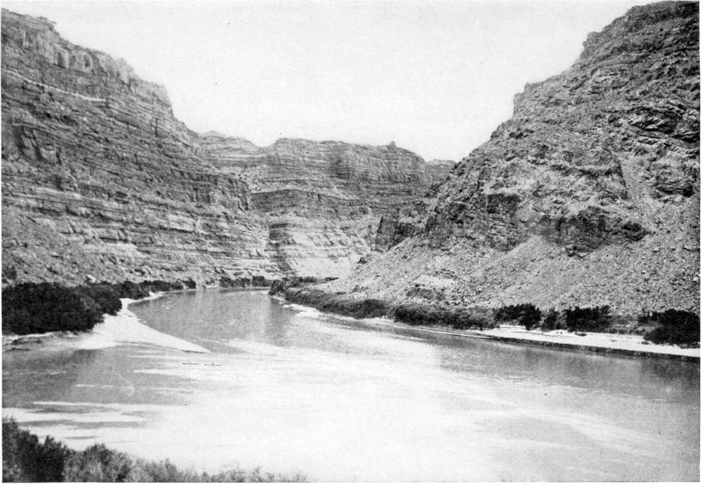 The river fills the bottom third of the image, curving between two low canyon walls. The mouth of another river joins at the lower left corner. On the lower right is a long, narrow sand beach lined with trees. Two boats are tiny dots on this beach.