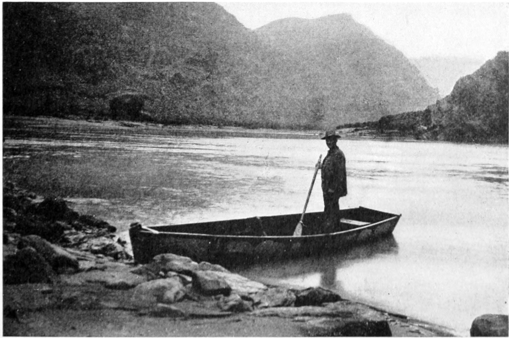 A man, holding an oar, stands at the stern of an open rowboat. The prow of the boat rests on a rocky shoreline. The placid river fills the lower half of the photo. Hills disappear into the distant horizon.