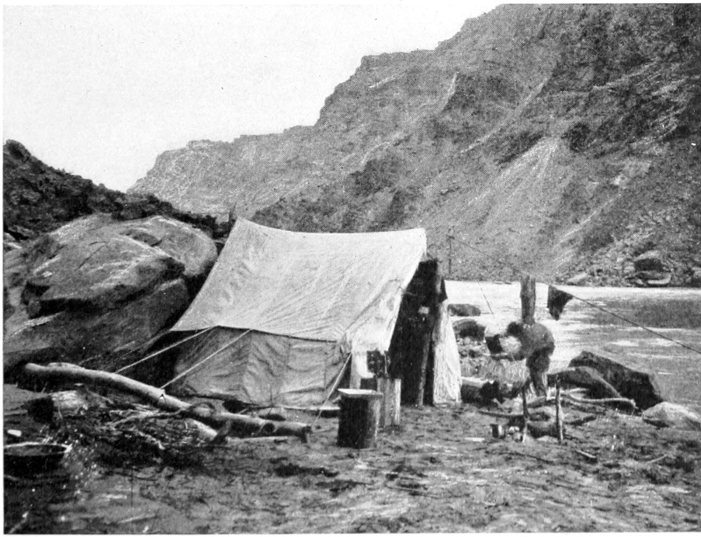 A tent is erected between large boulders and a flat piece of shoreline. Near the entry to the tent, a man holds a photographic plate between his hands. Pieces of cloth hang on a guy-wire from the top of the tent.