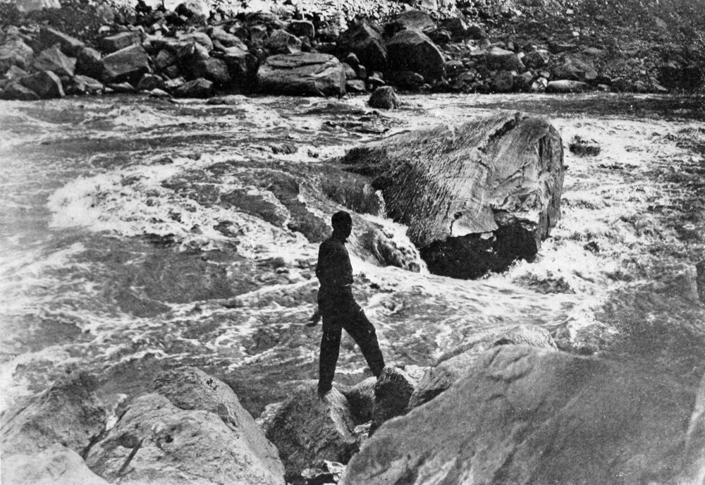 A man stands on large rocks next to the river. A huge boulder and other rocks fill the river. The water rushes through a chute, dropping perhaps ten feet.