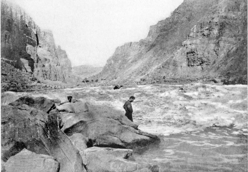A man stands on a large rock at the shoreline, watching torrents of water flow downstream. Canyon walls rise on both sides of the river.