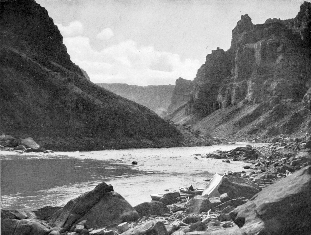 The Kolb boats are tied to a rocky shore in the canyon. A man is in one of the boats. Strewn among the large rocks are remnants of equipment washed up or left behind by a prior explorer.
