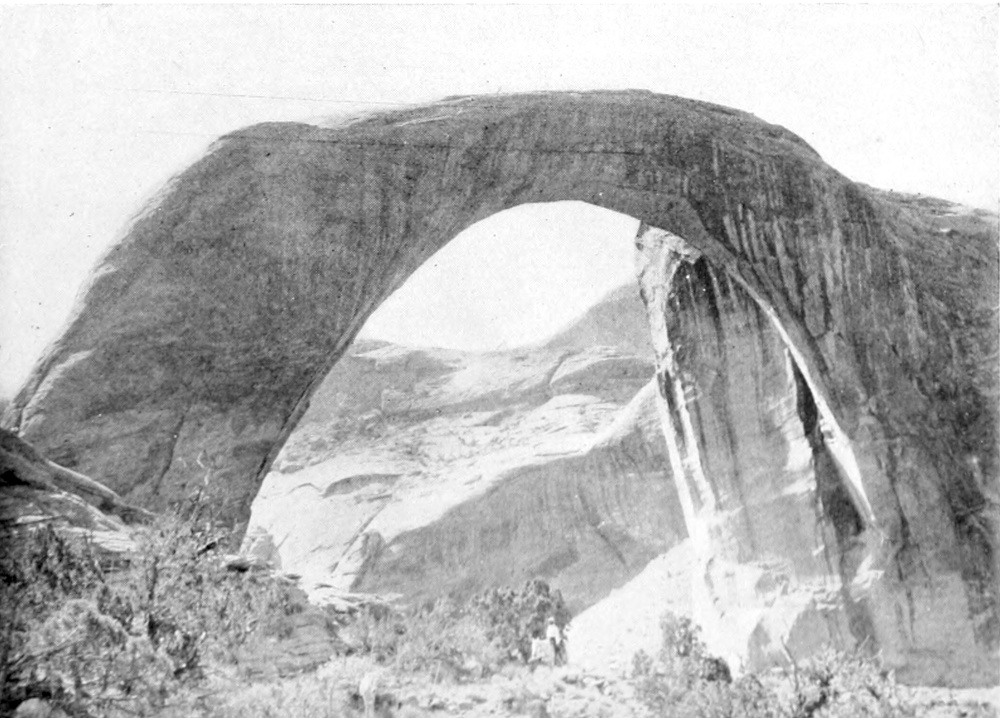 A natural stone arch spans a section of rocky canyon. The face of the bridge is in shadows; the canyon walls seen through the arch are sunlit.