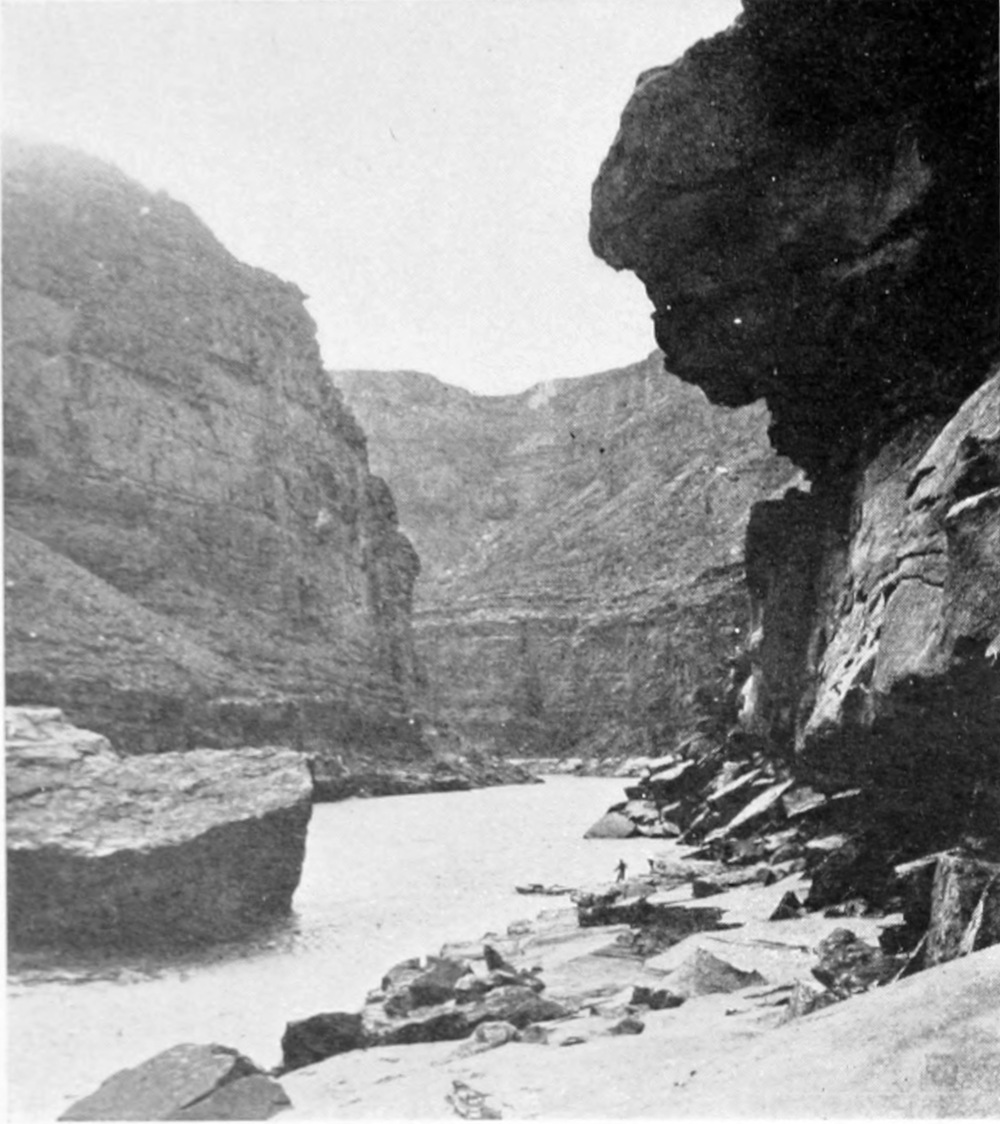 A rock wall overhangs the river. Rocks of various sizes line the sides of the river. A vertical canyon wall is opposite the overhang.