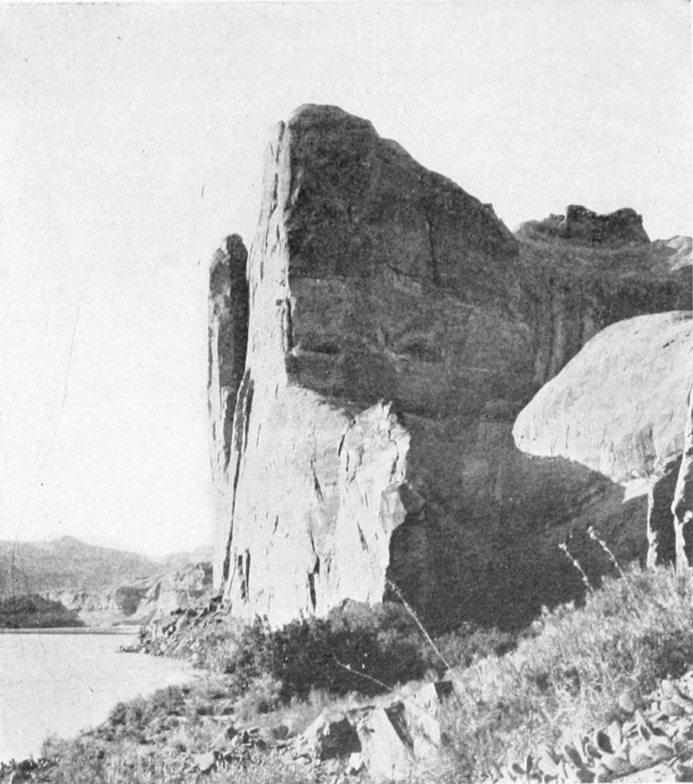 A sheer canyon face meets the river. Low hills are in the distance on the opposite side of the river.