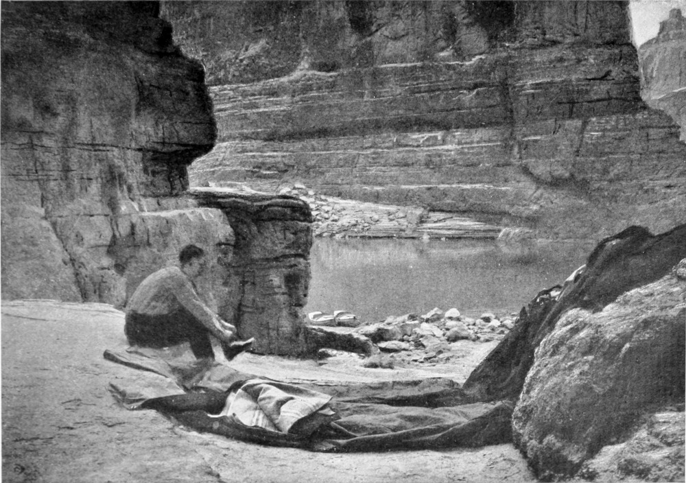 A man seated on a rock on shore ties his shoe. Sleeping bags are on the ground next to him. The two boats are grounded at the shoreline. A canyon wall rises above the opposite side of the river.
