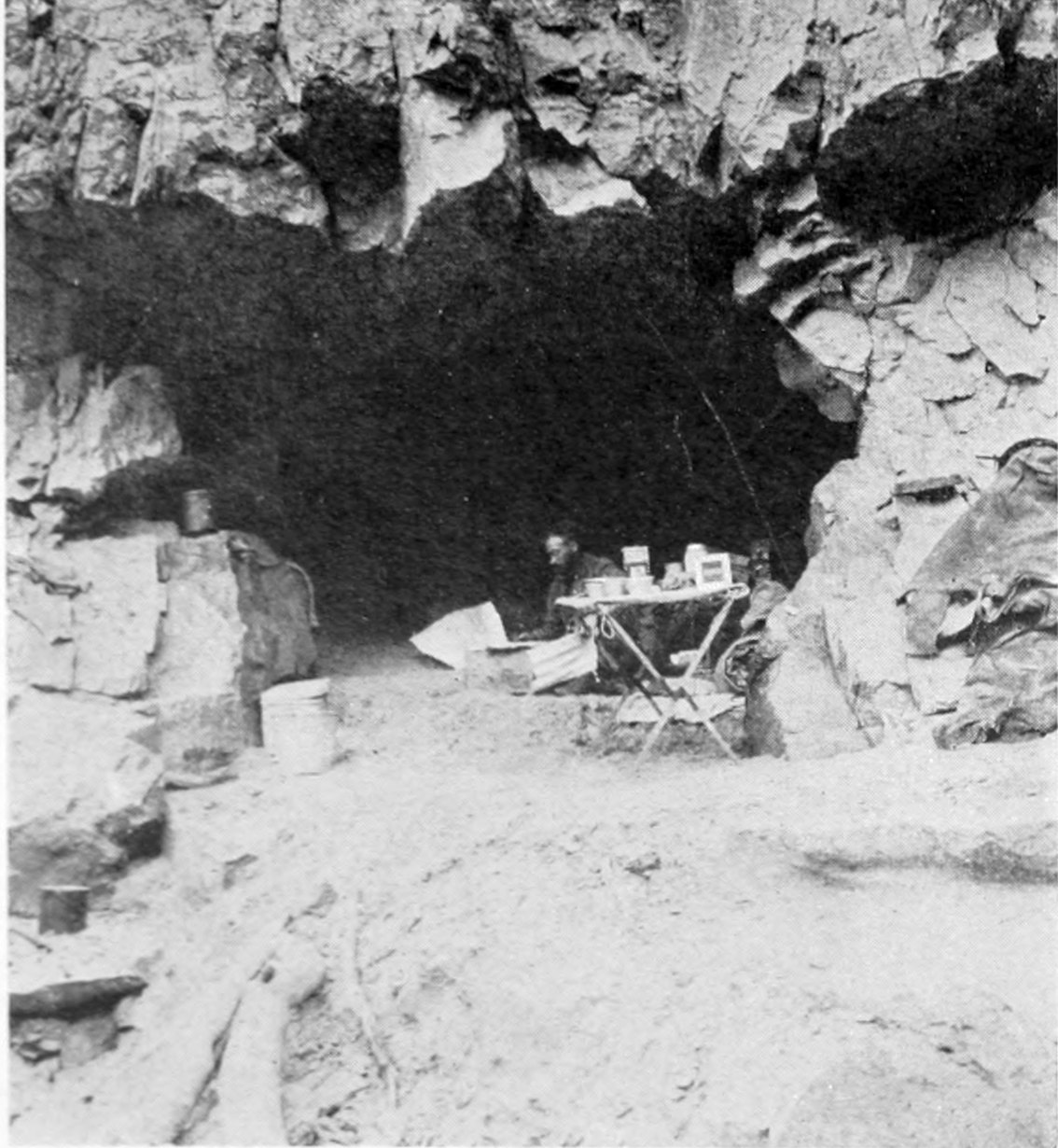 A collapsible table holds gear at the front of a cave in the canyon wall. A man is seated in the cave opening looking at a large sheet of paper (possibly a map).