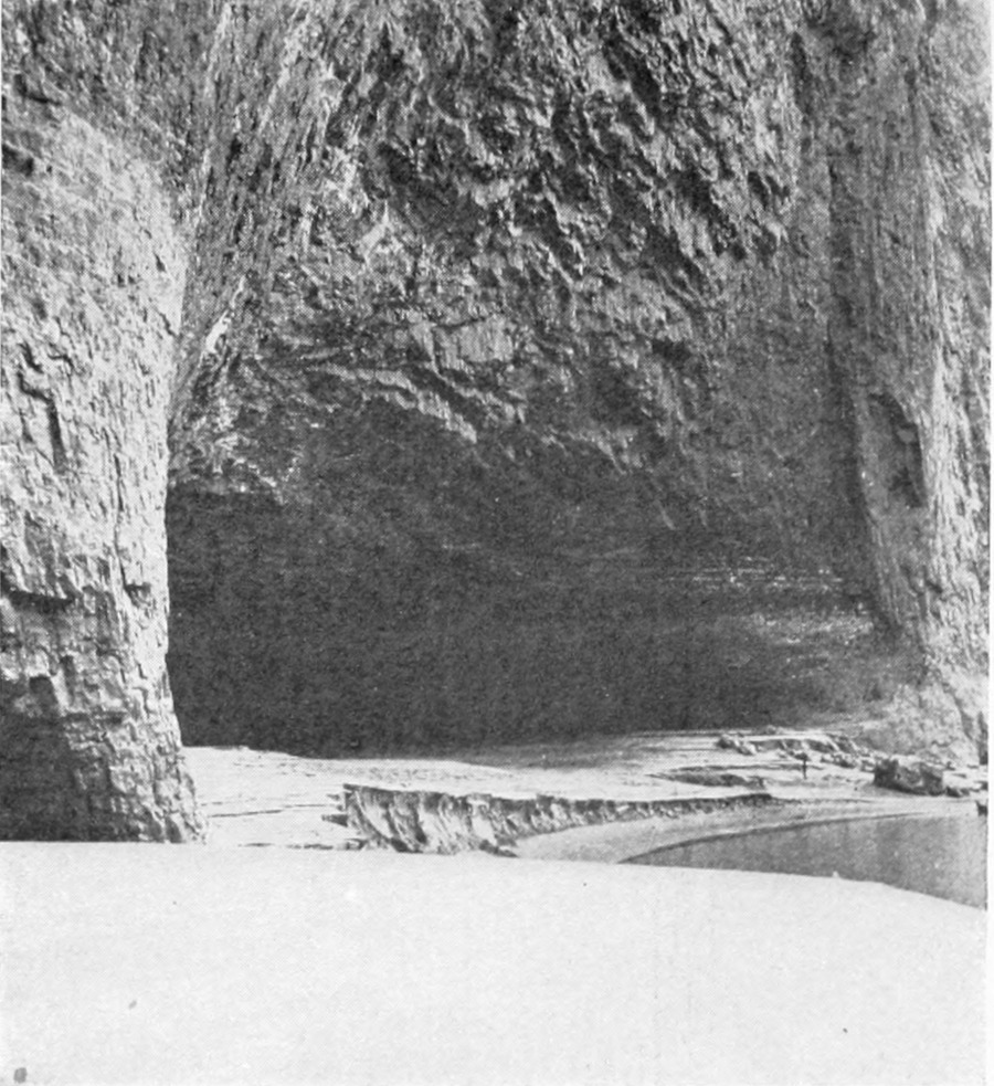 Before a cavernous opening into the canyon wall, a man at the shoreline is dwarfed by the opening.