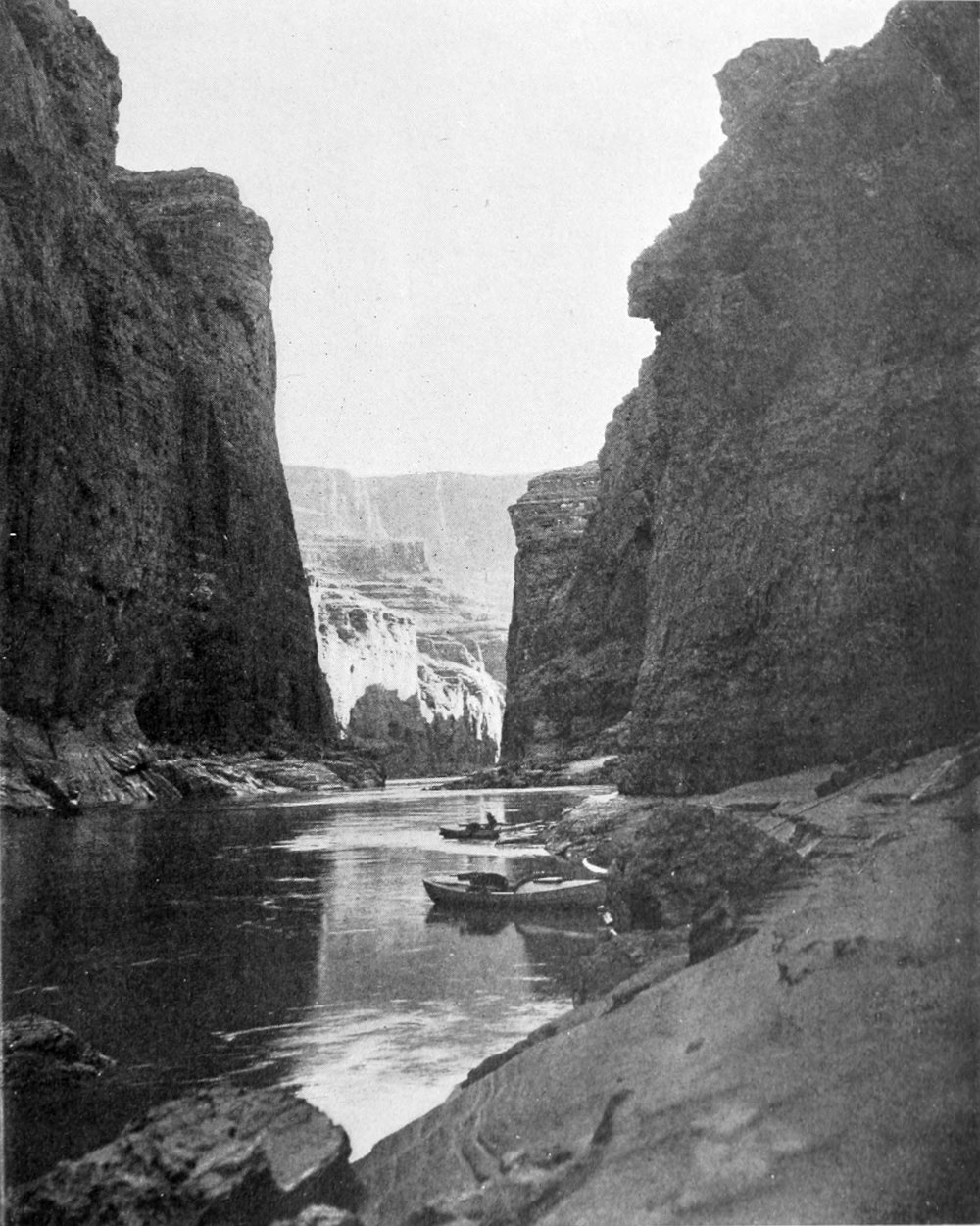 Two boats are tied at the shoreline. The canyon walls are close together, shaded from sunlight.