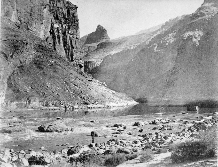 A man, standing on a rocky shore, looks across the river at a sloping canyon wall.