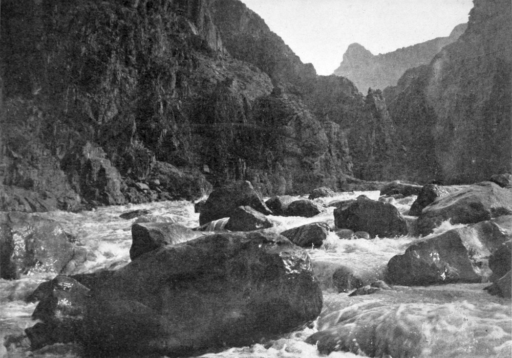Huge boulders fill the river, churning the water; a tiny boat in the distance prepares to run the rapids through the boulders.