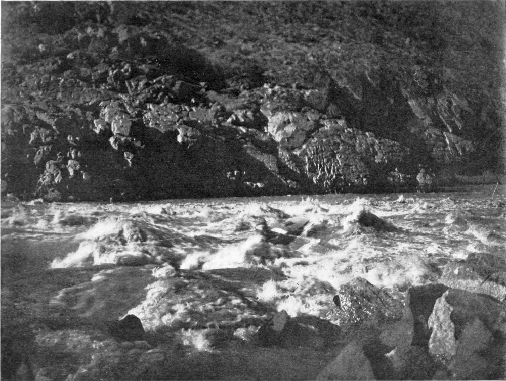 Water rushes over rocks across the width of the river. A rocky hillside slopes away from the far side of the river.