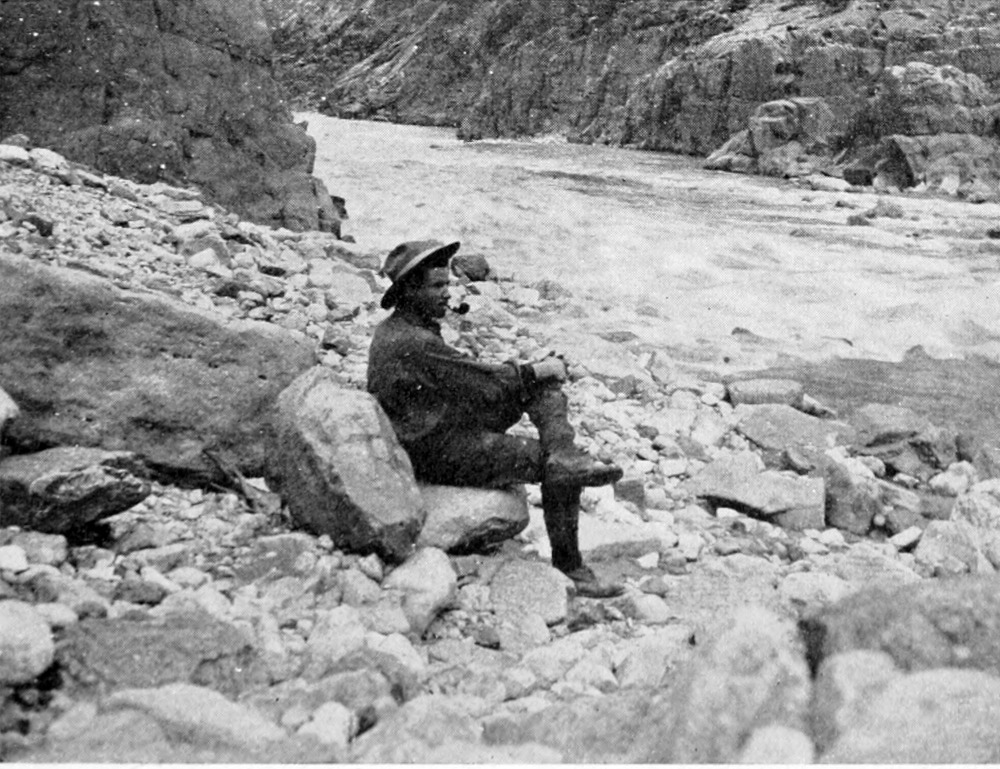 Lauzon sits, smoking a pipe, with one leg crossed over the other on a rock at the shoreline of the river.