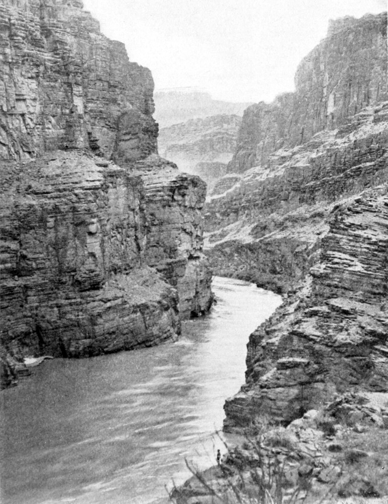 The river passes between two high canyon walls. There is no shore on either side. A tiny figure of a man is standing at the bottom of the picture, providing a sense of scale of the height of the canyon.