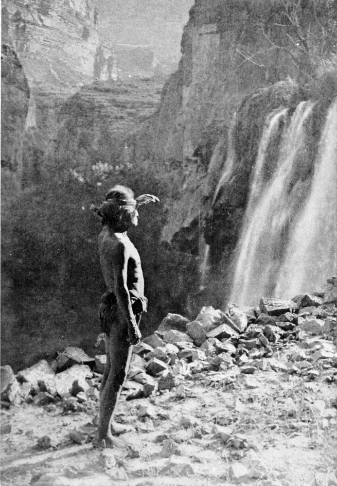 A barefoot man wearing a simple feather headdress and a loincloth stands in the sunlight facing a waterfall.
