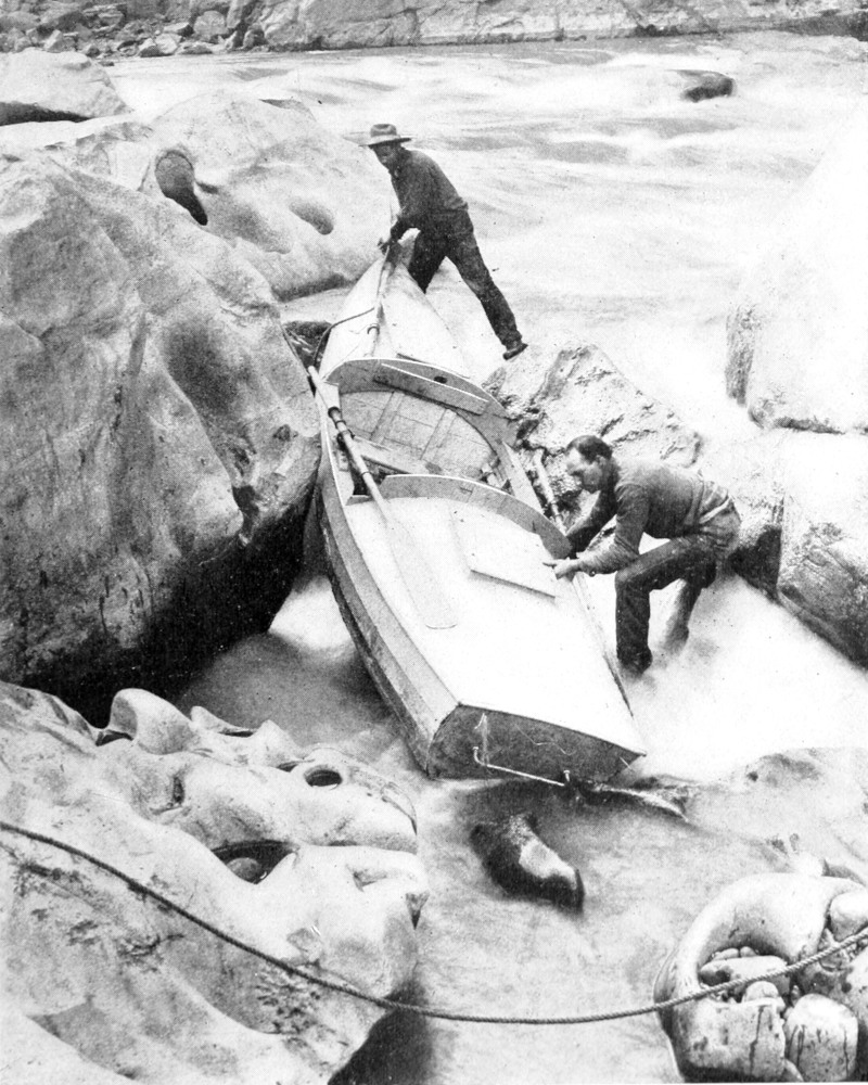 Two men lifting a boat around huge boulders next to the river.
