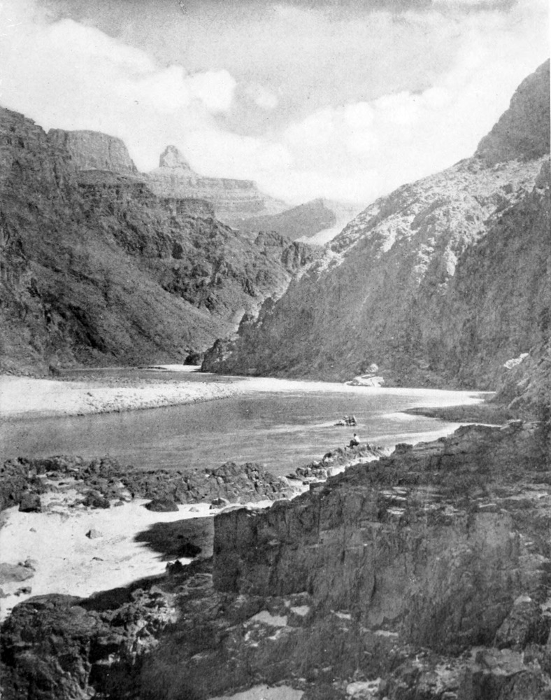 A man sits on rocks at the river shore. Canyon walls rise on all sides.