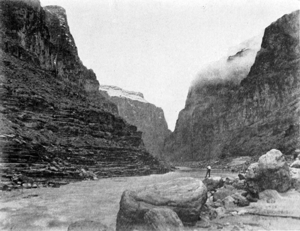 A man stands on rocks next to the river. Clouds obscure the upper canyon walls.