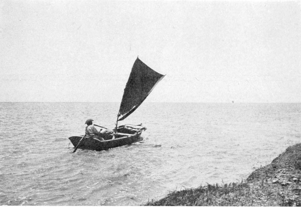 Man rowing a boat with a triangular sail on a wide expanse of water.